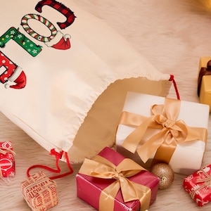 May include: A cream-colored Christmas gift bag with the word "ELF" in festive lettering. The bag is open, revealing wrapped gifts in red, white, and gold, with gold ribbons and bows. Christmas ornaments and candles are also visible.