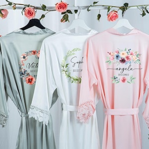 May include: Three satin bridesmaid robes in sage green, white, and pink, hanging on a rack decorated with roses. Each robe has a floral design and a name printed on the back. The robes have lace trim on the sleeves.