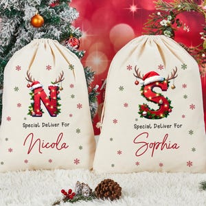 Puede incluir: Dos bolsas de regalo navideñas color crema. Una bolsa presenta la letra roja "N" decorada con luces, cuernos y un gorro de Papá Noel, con el nombre "Nicola" debajo. La otra bolsa tiene una "S" roja con el nombre "Sophia".