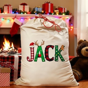 May include: A large, off-white Christmas sack with the name "JACK" in festive lettering. The letters are decorated with a Santa hat, antlers, and a gingerbread man. A red drawstring and a teddy bear are also visible.