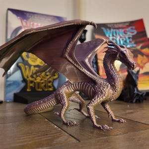 May include: A purple and gold dragon figurine with wings spread wide. The dragon is posed with its head turned to the side and its mouth slightly open. The figurine is made of plastic and is about 10 inches long.