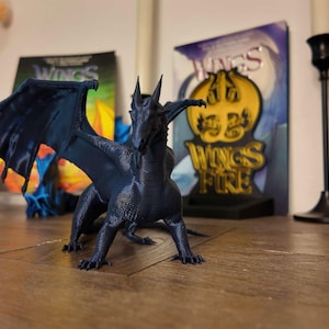 May include: A blue 3D printed dragon figurine with wings outstretched. The dragon is posed in a standing position with its head tilted slightly to the side. The figurine is set against a background of books with the title "Wings of Fire" visible.