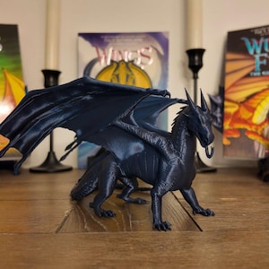 May include: A dark blue 3D printed dragon figurine with wings spread wide. The dragon is posed with its head tilted slightly to the side and its mouth open. The figurine is sitting on a wooden table in front of three books with the title "Wings of Fire" on the covers.