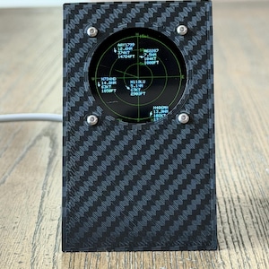 Könnte beinhalten: Ein rechteckiges elektronisches Gerät mit einem schwarz-grauen Kohlefaser-Muster. Es verfügt über ein kreisförmiges digitales Display, das grüne Radar-Flugdaten mit Text wie AAY1799, N62287, N734HD, N118LU und N406MA anzeigt und mit vier silbernen Schrauben befestigt ist.