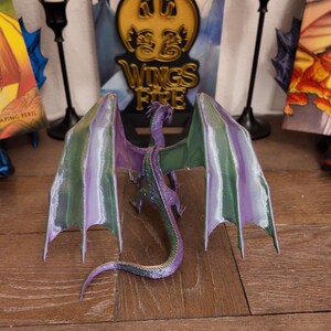 May include: A purple and green dragon figurine with wings spread wide. The dragon is posed in a flying position with its head tilted back. The figurine is made of plastic and has a detailed paint job. The dragon is sitting on a wooden surface. The words "Wings of Fire" are visible in the background.