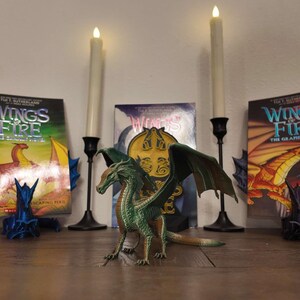 May include: A green dragon figurine with wings spread wide, posed in front of three books titled "Wings of Fire" with a dragon design on the cover. The books are on a table with two lit candles on either side.