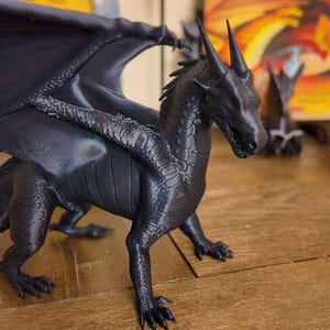 May include: A black 3D printed dragon figurine with wings outstretched. The dragon has a detailed textured surface and is posed in a standing position.