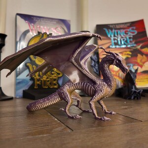 May include: A purple and gold dragon figurine with wings spread wide. The dragon is posed in a standing position with its head turned to the side. The figurine is sitting on a wooden table with three books in the background. The books are all titled "Wings of Fire".