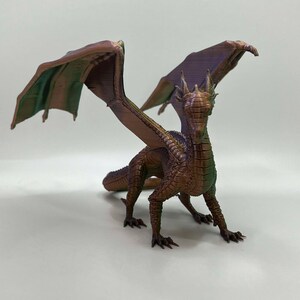 May include: A 3D printed dragon figurine with iridescent bronze and green wings. The dragon is standing on a white surface with its wings spread out.
