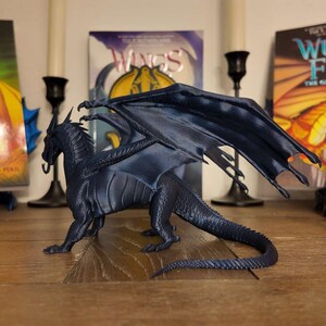 May include: A dark blue dragon figurine with wings outstretched, posed on a wooden surface. The dragon is a popular character from the Wings of Fire book series.
