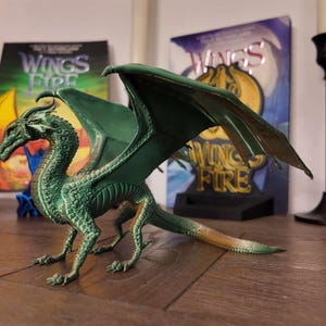 May include: A green dragon figurine with wings outstretched, standing on a wooden surface. The dragon has a detailed, textured body and a gold-colored underbelly. The figurine is surrounded by books with the title "Wings of Fire" on the cover.