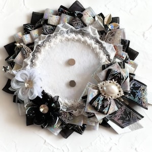 Könnte beinhalten: Dekorative, kreisförmige Brosche mit einem weißen Filzmittelteil und zwei silbernen Magneten. Die Brosche ist mit schwarzen, weißen und silbernen Bändern, Spitze und floralen Akzenten verziert. Eine kleine Sukkulentenpflanze in einem weißen Topf befindet sich im Hintergrund.