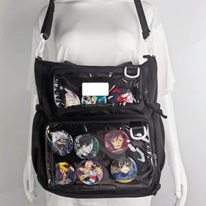 Könnte beinhalten: Schwarze Umhängetasche mit transparenten Vordertaschen, die bunte Anime-Charakter-Buttons zeigen. Die Tasche hat einen langen, verstellbaren Riemen und einen weißen Verschluss. Die Tasche wird auf einer weißen Schaufensterpuppe präsentiert.
