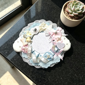 Può includere: Una cornice fotografica circolare decorativa con un centro bianco, circondata da nastri azzurri e rosa, pizzo e piccoli ciondoli a forma di croce. La cornice include un abbellimento a forma di cuore e il testo "Home Home". Una pianta succulenta in un piccolo vaso è sullo sfondo.