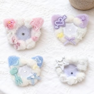 May include: Four pastel-coloured plush cat ear hair clips. Each clip has a different design, including bows, stars, and floral accents. The clips are arranged on a white, textured surface, with a small potted succulent in the background.