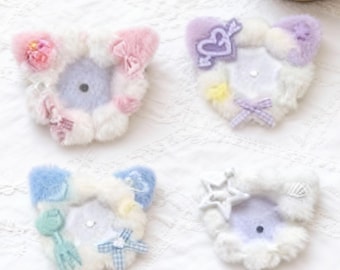 May include: Four plush cat ear hair clips in pastel colors. Each clip features a different design, including bows, stars, and floral accents. The clips are arranged on a white, textured surface, with a small potted succulent in the background.