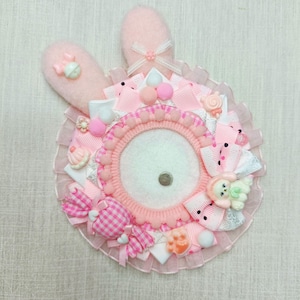 May include: A decorative bunny-shaped accessory in pink and white. The design includes a central white circle, surrounded by pink ribbons, bows, and small embellishments. The bunny ears are pink, with a small bow at the base of one ear.