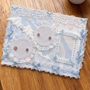 May include: A decorative piece in light blue and white, featuring lace trim, bows, and felt bat and circle details. It includes a frame and two felt circles with magnets. A small succulent plant in a white pot is in the background.
