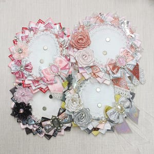 May include: Four decorative, circular brooches with white centers and ornate ribbon and floral embellishments. The brooches feature pink, silver, black, and gray ribbons, bows, and fabric flowers. Each brooch has a magnetic closure.