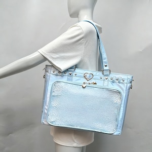 May include: Pale blue tote bag with a clear front pocket and silver-tone hardware. The bag has a heart-shaped clasp and decorative studs. The handles are also pale blue. The bag is held by a mannequin.