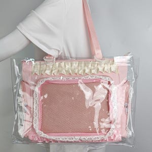 K&ouml;nnte beinhalten: Eine rosa Haustiertasche mit transparentem Kunststoff au&szlig;en. Die Tasche hat ein Netzfenster, Spitzenbesatz und einen rosa Schultergurt. Die Tasche hat eine dekorative R&uuml;sche am oberen Rand und zwei Seitentaschen mit Rei&szlig;verschluss.