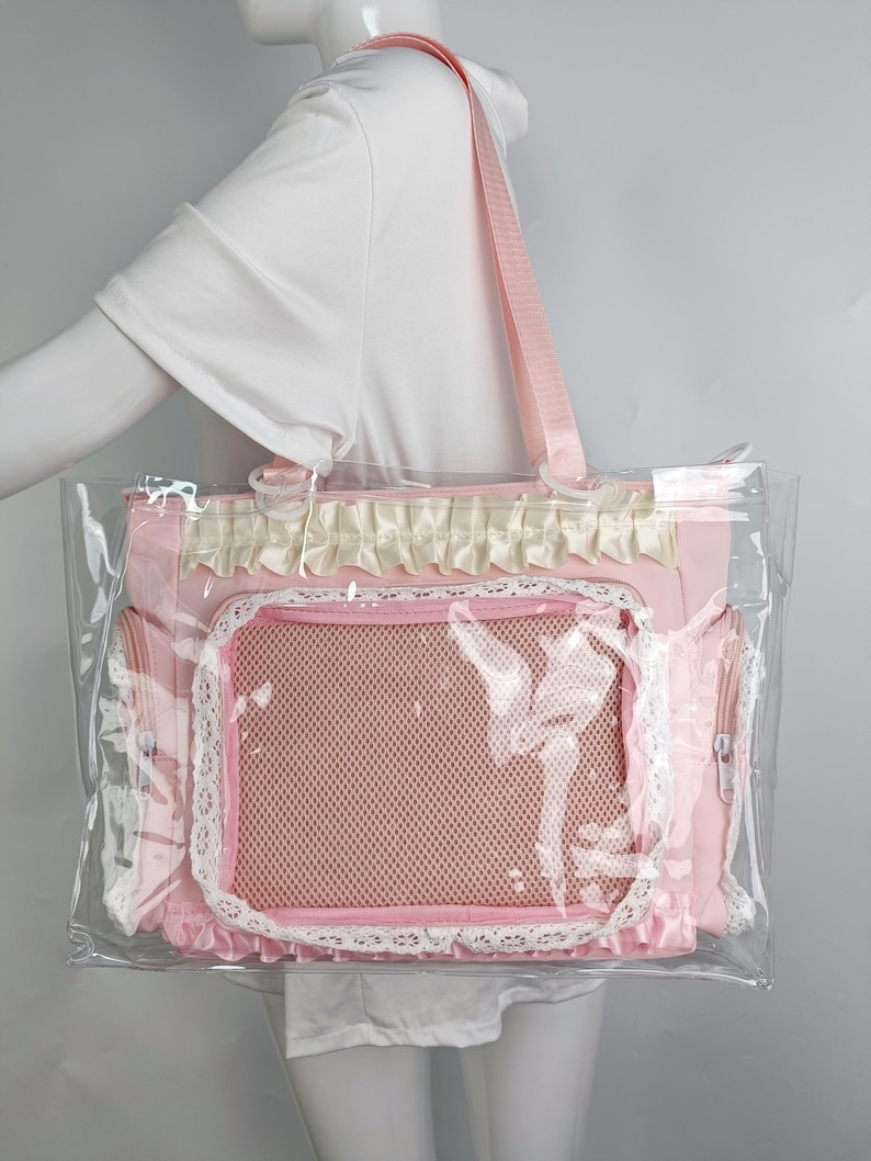 K&ouml;nnte beinhalten: Eine rosa Haustiertasche mit transparentem Kunststoff au&szlig;en. Die Tasche hat ein Netzfenster, Spitzenbesatz und einen rosa Schultergurt. Die Tasche hat eine dekorative R&uuml;sche am oberen Rand und zwei Seitentaschen mit Rei&szlig;verschluss.