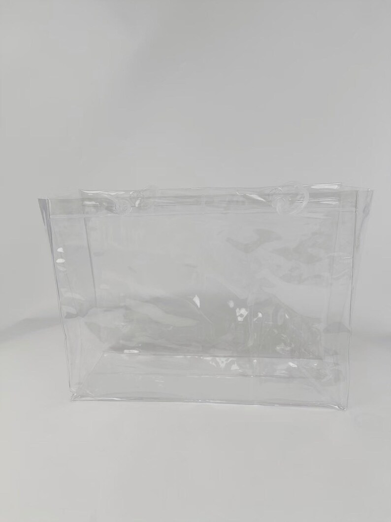 K&ouml;nnte beinhalten: Eine transparente Tragetasche mit zwei Griffen. Die Tasche besteht aus einem flexiblen, durchsichtigen Material und hat eine rechteckige Form. Die Tasche ist leer und steht vor einem wei&szlig;en Hintergrund.