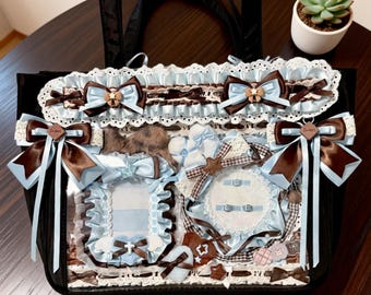 May include: Black tote bag with decorative elements. Features light blue and brown bows, lace trim, and decorative frames. The bag has a clear front panel displaying the interior details. A small succulent plant in a white pot is in the background.