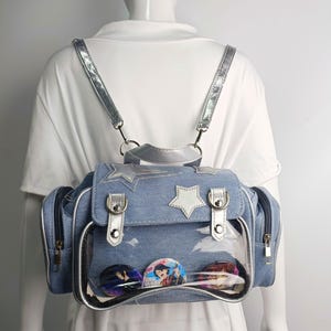 May include: Light blue denim backpack with silver accents, including straps, buckles, and star-shaped embellishments. Features a clear front pocket displaying colorful pinback buttons. Includes two side pockets with zippers.