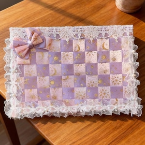 Puede incluir: Una alfombra rectangular decorativa con un patrón de tablero de ajedrez tejido en tonos lavanda y rosa, adornada con diseños celestiales dorados. La alfombra está adornada con encaje blanco y un lazo rosa.
