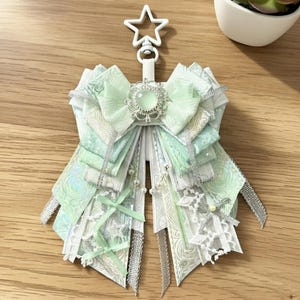 May include: A decorative bow-shaped keychain in shades of mint green and white, adorned with silver accents and a star-shaped clasp. The bow features layered ribbons, lace, and a central jewel. A small succulent plant is in the background.