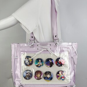 K&ouml;nnte beinhalten: Eine lavendelfarbene Tragetasche mit einem transparenten Frontpanel, das acht farbenfrohe Charakterpins zeigt. Die Tasche hat einen passenden lavendelfarbenen Riemen und silberfarbene Hardware. Die Tasche besteht aus einem klaren, transparenten Material.