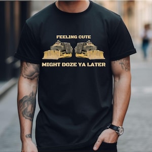Może przedstawiać: Czarny T-shirt z grafiką dwóch żółtych buldożerów zwróconych do siebie. Tekst "Feeling Cute" znajduje się nad buldożerami, a "Might Doze Ya Later" pod nimi.
