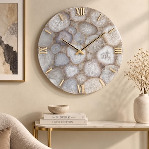 May include: A round wall clock featuring a face made of multi-toned beige and gray agate slices. The clock has gold-colored Roman numerals at the twelve, three, six, and nine o'clock positions, with thin gold-colored hands and simple gold-colored markers.