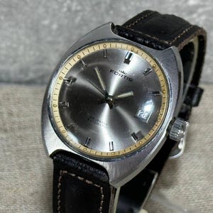 May include: A silver wristwatch with a black leather strap. The watch face is silver with gold accents and the word "FORTIS" printed on it. The watch has a date window at the 3 o'clock position.