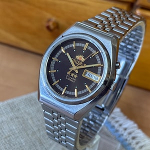 Può includere: Un orologio da polso vintage Orient Crystal 21-Jewels con cinturino in acciaio inossidabile color argento. Il quadrante dell'orologio è nero con lancette e indici dorati e mostra il giorno e la data. L'orologio è impostato su venerdì 18.