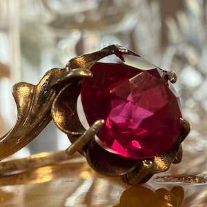May include: A vintage gold ring with a large, oval-cut ruby gemstone. The ring has an ornate, floral design. The ruby is a deep red colour, and the ring is set against a blurred background. The ring's design is reminiscent of a flower.