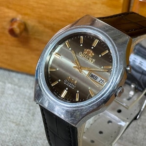May include: An Orient wristwatch with a silver-tone metal case, a brown sunburst dial, and gold-tone hour markers and hands. The watch features a day-date window at the three o'clock position and a black leather strap with a textured finish.