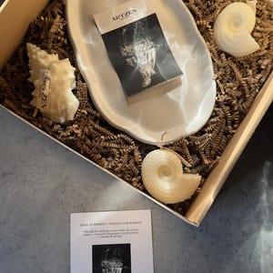 Puede incluir: Una caja de regalo que contiene un plato de cerámica blanco de forma ovalada, tres velas con forma de concha y una tarjeta con el texto "ARTZEN DECOR". La caja está llena de papel arrugado marrón.