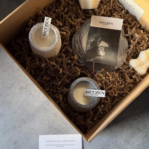 May include: A gift box containing various decorative candles and a card. The box includes two small, stone-effect candles, a large wrapped candle, and a white, textured candle. The card displays the text "ARTZEN DECOR".