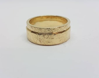 Könnte beinhalten: Zwei goldene Eheringe, die übereinander liegen. Die Ringe haben eine strukturierte Oberfläche.