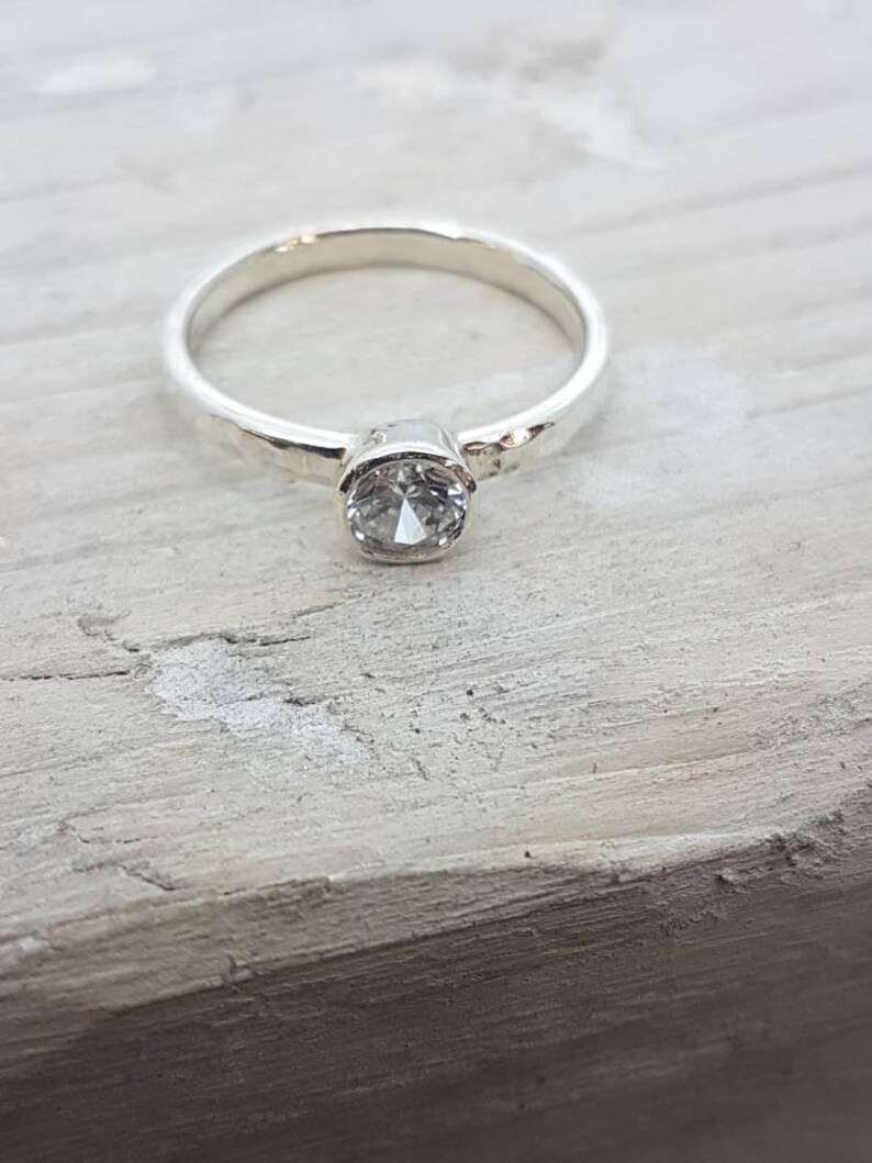 K&ouml;nnte beinhalten: Ein silberner Ring mit einem geh&auml;mmerten Band und einem klaren, runden Edelstein.