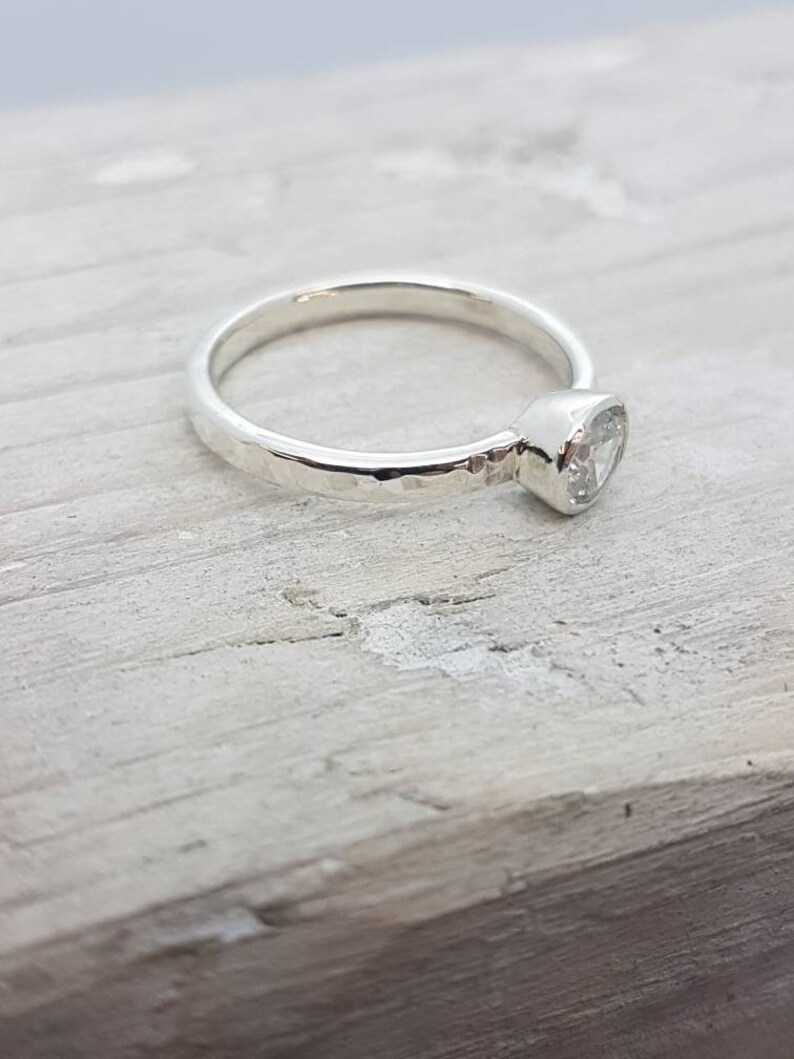 K&ouml;nnte beinhalten: Ein silberner Ring mit einem geh&auml;mmerten Band und einem gefassten, klaren Edelstein.