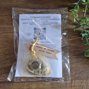 Op de afbeelding: Een doorzichtige plastic zak met een charmpocket met de tekst "Pearls of Wisdom Charm Pocket". De pocket is vastgebonden met een geel lint en bevat een kleine hanger en gedroogde kruiden. Een witte kaart met tekst en een QR-code is zichtbaar.