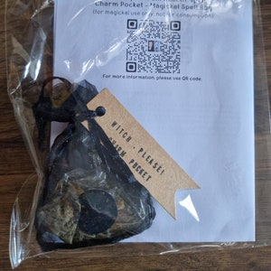 May include: A black mesh charm pocket with dried herbs and a black stone, tied with a black ribbon and a brown tag that reads "Witch, Please! Charm Pocket." A white paper with text and QR codes is behind the charm pocket.