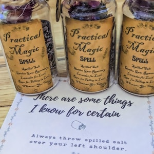 Puede incluir: Tres frascos de vidrio transparente rellenos de hierbas secas y flores moradas, cada uno con una etiqueta color canela con el texto Practical Magic SPELL. Debajo hay una tarjeta blanca con el texto There are some things I know for certain.