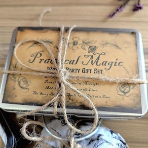May include: A rectangular metal tin with a tan label featuring the text Practical Magic PARTY GIFT SET and floral illustrations, tied with brown twine. Below the tin is a white fabric pouch with a black botanical print, also tied with twine.