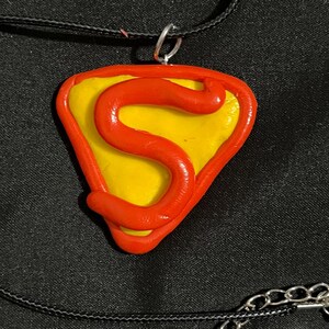 May include: A handmade necklace featuring a pendant with a yellow center and a red outline shaped like a shield. A red letter "S" is in the center. The pendant hangs from a black cord with a silver-colored clasp.
