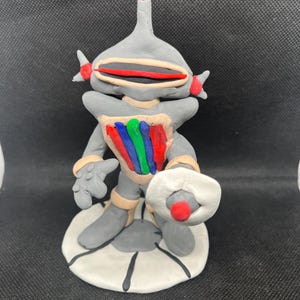 Puede incluir: Figura de robot de arcilla gris hecha a mano con detalles en rojo, azul, verde y beige. El robot tiene una antena roja, una boca roja y beige, y un objeto blanco en forma de disco con un punto rojo. Se encuentra sobre una base blanca.