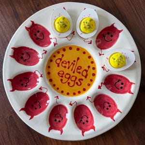 May include: A round white ceramic serving platter for deviled eggs featuring a central yellow circle with the text deviled eggs in red. The platter has twelve red painted devil characters with horns and tails, and three egg halves with yellow filling.